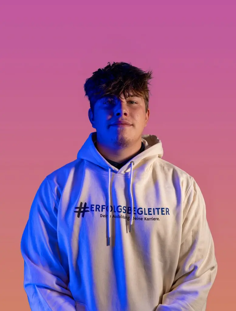 Malte_Baerlocher Startseite: Malte Baerlocher im Hoodie mit #ERFOLGSBEGLEITER-Aufdruck. Porträt vor farbigem Hintergrund.