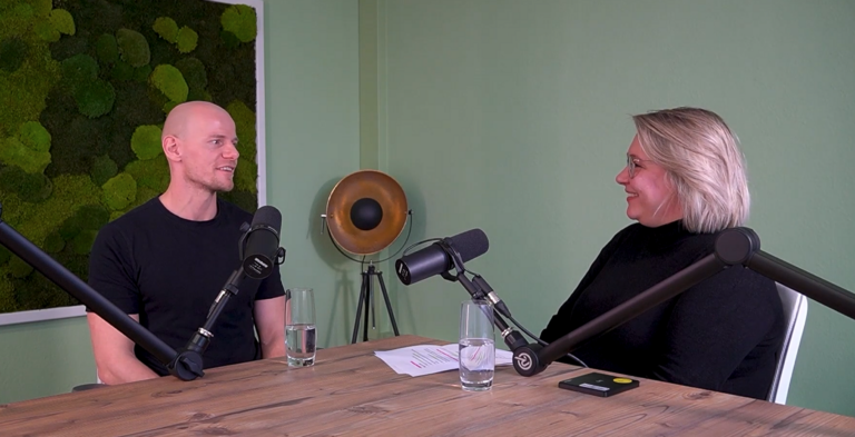 Podcastaufnahme: Zwei Personen mit Mikrofonen am Tisch, im Hintergrund grüne Wand. Thema: Glücklich im Job und in der Ausbildung, Zufriedenheit.